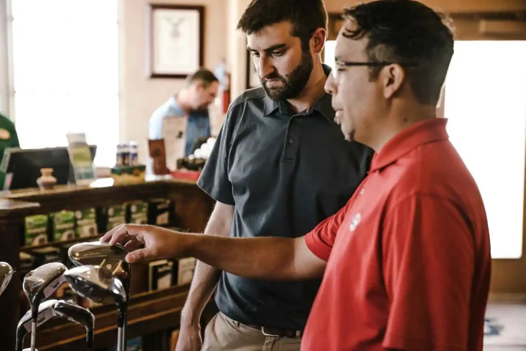 Two men discussing and examining putter grips in a pro shop, aiming to select the best golf grips for arthritis.