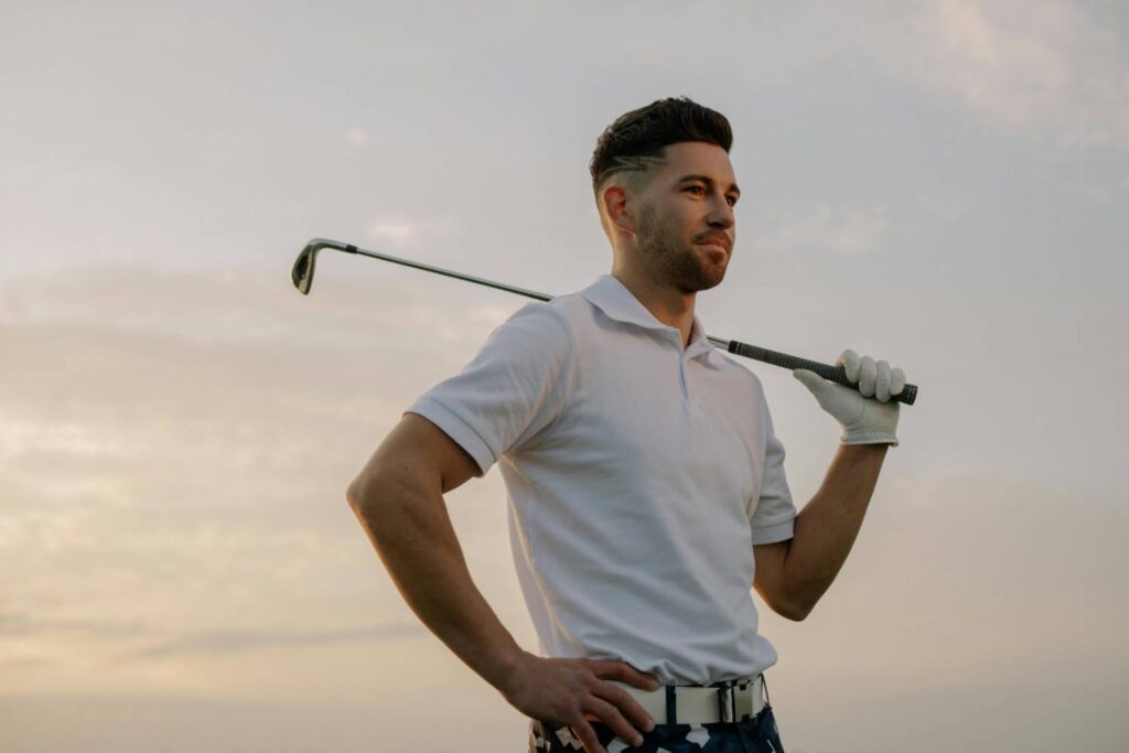 A confident golfer holding a club over his shoulder, wearing a white polo shirt and golf gloves, with a scenic sunset background, hoding a putter grip