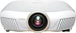 This image shows a white Epson Home Cinema projector. The lens is centrally located and surrounded by a gold-colored ring. The Epson logo is visible on the left side of the projector's front. Ventilation grilles are present on both the left and right sides.