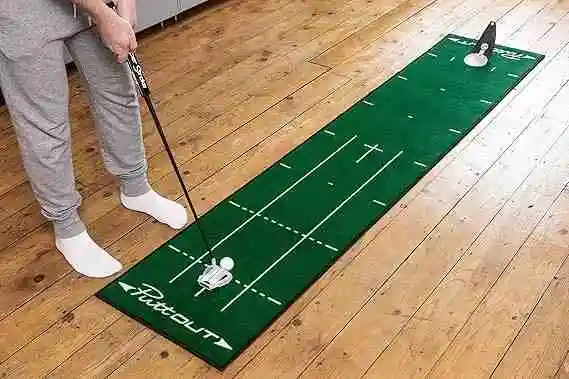 A person wearing grey sweatpants and white socks uses a putter on a green PuttOut putting mat with white alignment lines and a PuttOut pressure putt trainer at the far end.