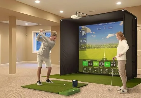 A man swings a golf club while using the SkyTrak+ Golf Simulator Package in a home setting, as a woman watches the simulated golf course displayed on a large screen.